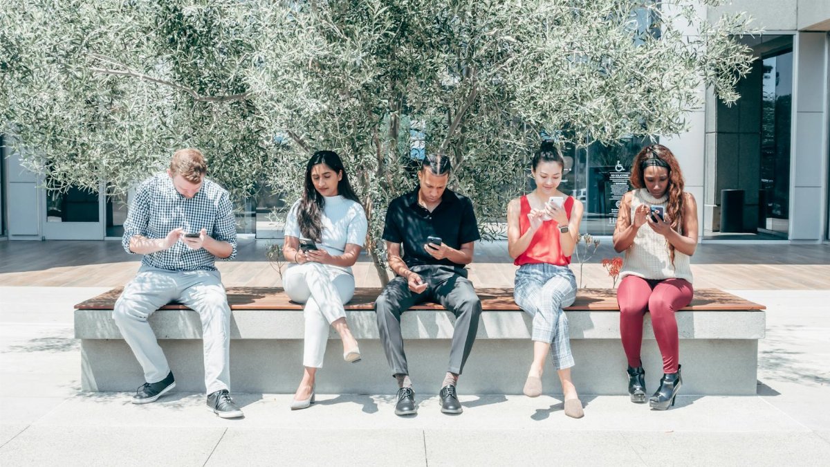 Five diverse people sitting on a bench outdoors, engaged with their smartphones.