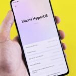 Close-up of a smartphone displaying Xiaomi HyperOS interface, held by a hand against a bright yellow background.