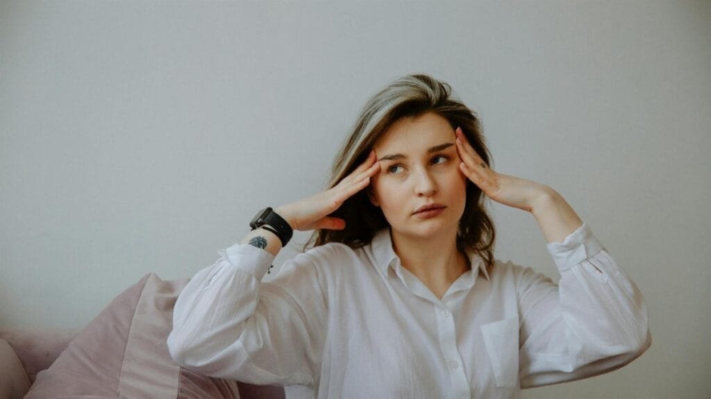 Woman in white shirt looking stressed, touching her temples indoors.
