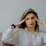 Woman in white shirt looking stressed, touching her temples indoors.