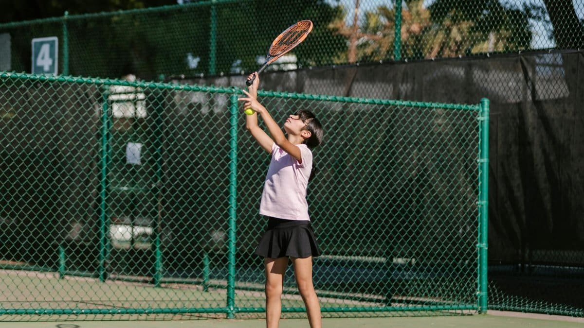 A young girl practices tennis, preparing to serve on an outdoor court.