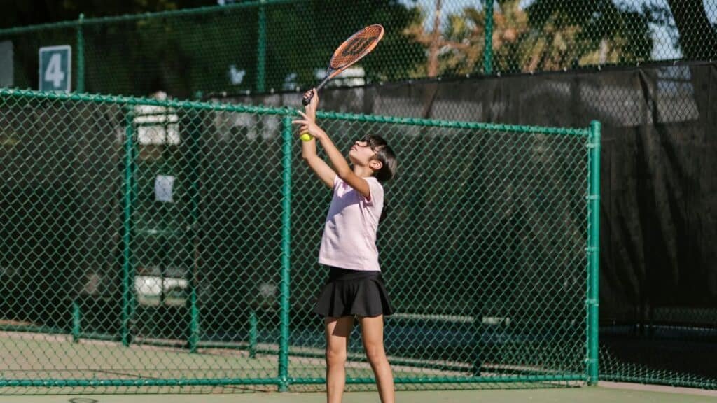 A young girl practices tennis, preparing to serve on an outdoor court.