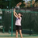 A young girl practices tennis, preparing to serve on an outdoor court.