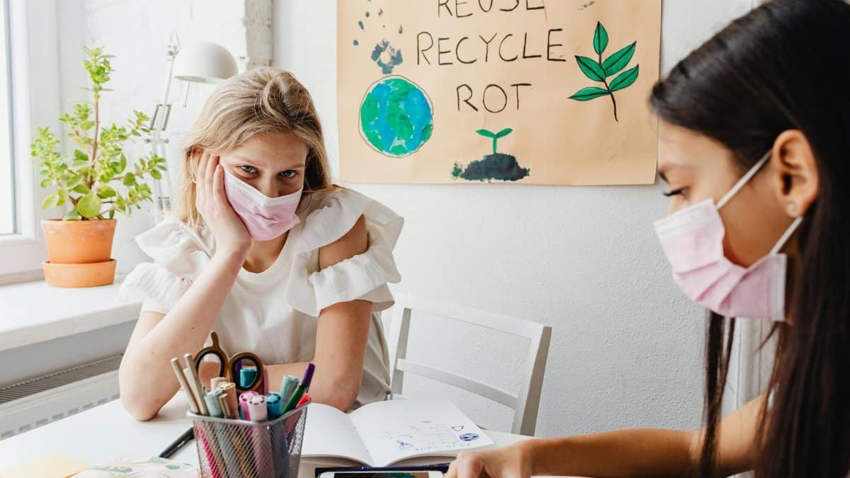 Two young women in masks studying with eco poster and plants nearby, promoting sustainability.