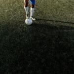 A soccer player stands on a field with a ball under bright sunlight, casting a long shadow.