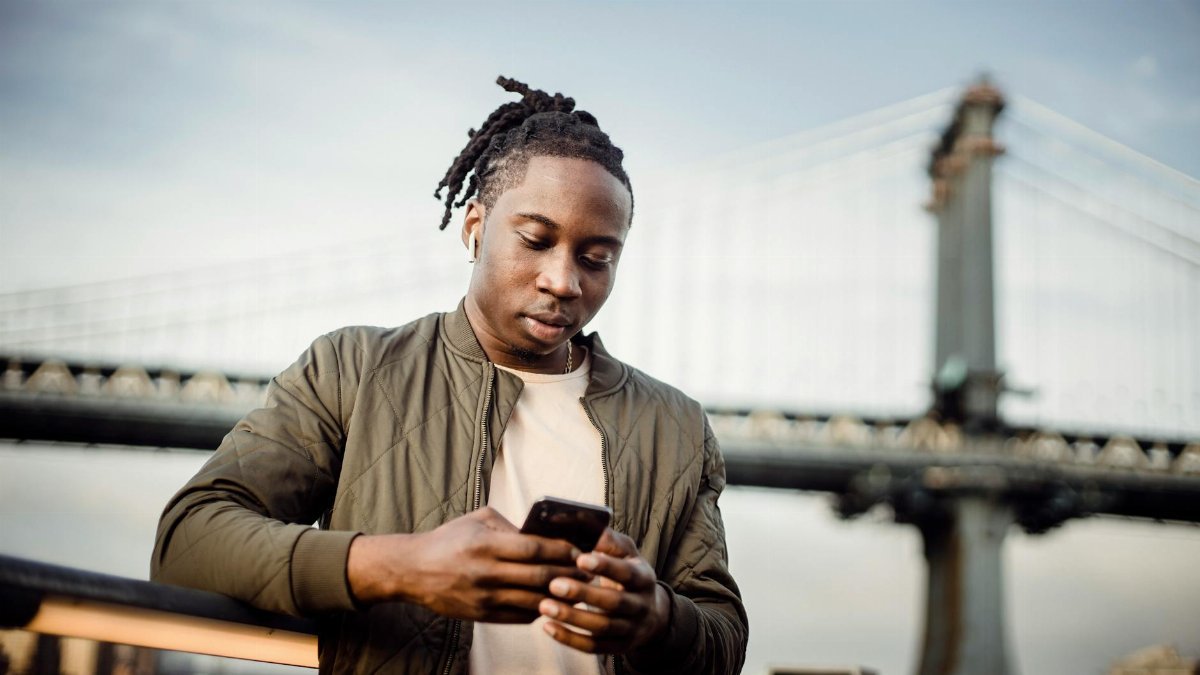 From below of serious black man in earphones scrolling smartphone while listening to music outside and looking down