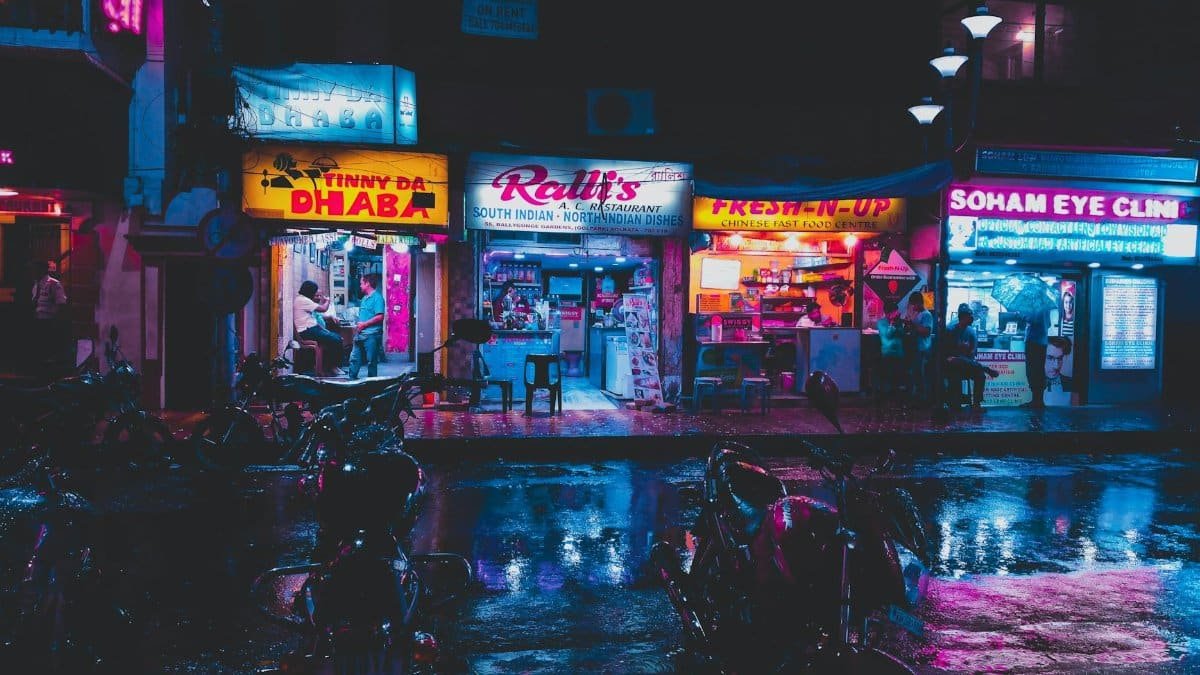 Colorful street scene in Kolkata with glowing shop signs and wet pavement at night.