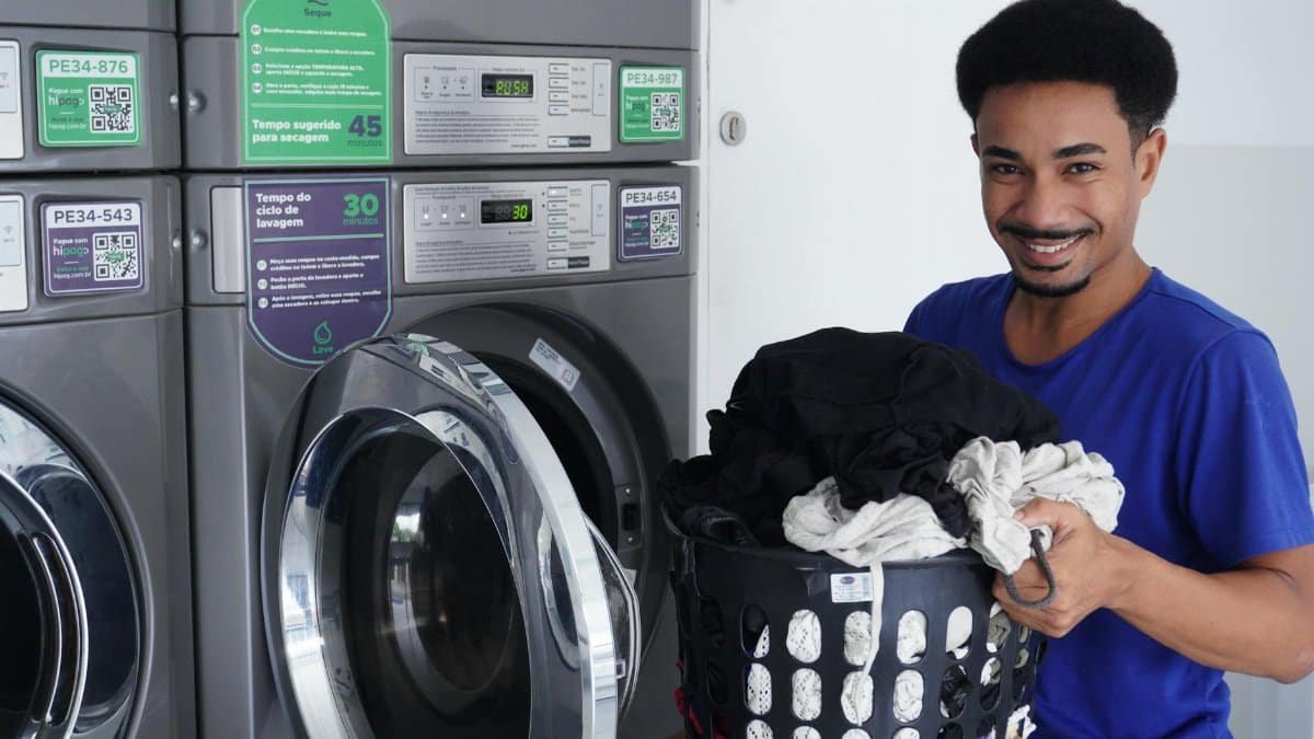 Smiling man holding laundry basket in a laundromat, ready to wash clothes.