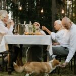 Seniors enjoying an outdoor dinner with a corgi under string lights in a forest setting.
