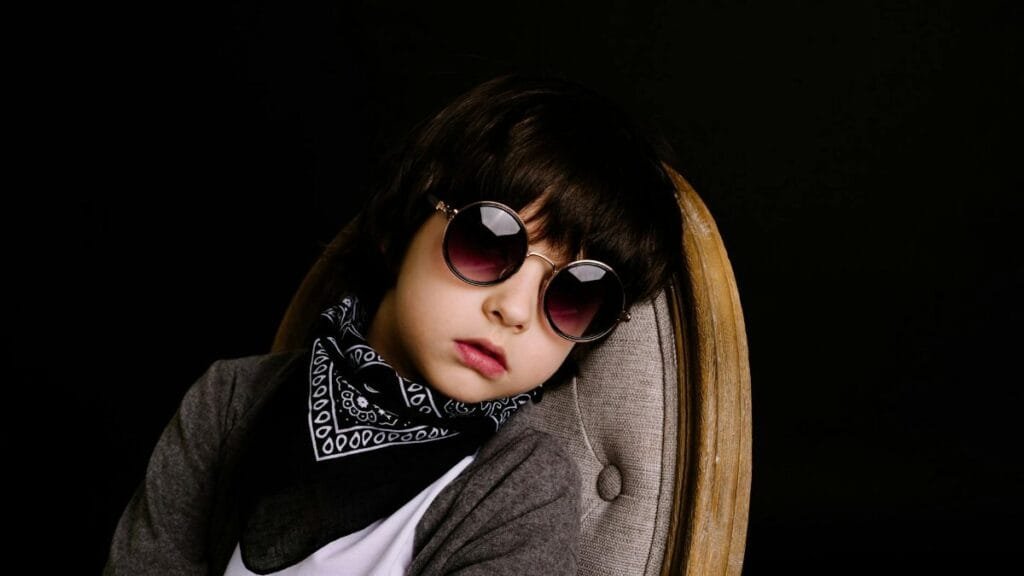 Portrait of a fashionable child wearing sunglasses and a scarf, sitting on a chair against a dark background.