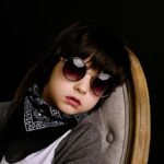 Portrait of a fashionable child wearing sunglasses and a scarf, sitting on a chair against a dark background.