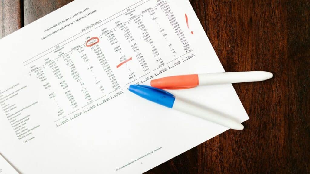 Top view of financial document with red marks and pens on wooden table surface.