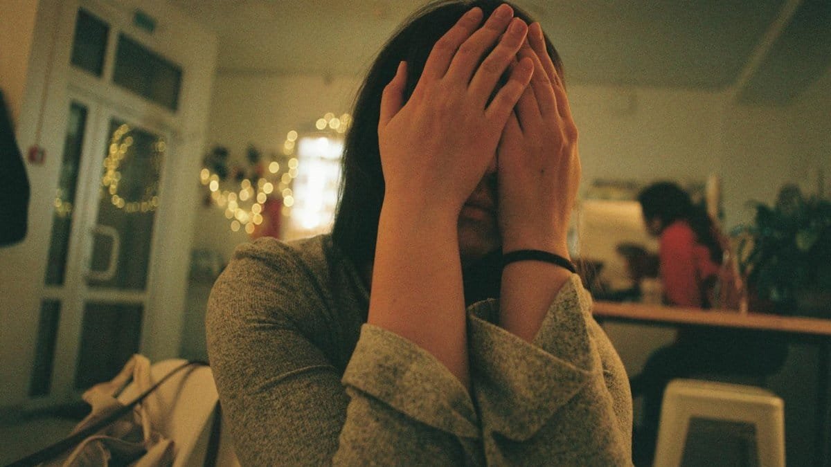 A woman covers her face with hands, expressing stress in a cozy room.