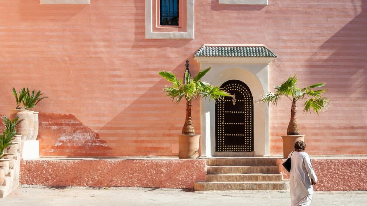 A Muslim woman walking past a traditional Moroccan building facade in Marrakesh during the day.