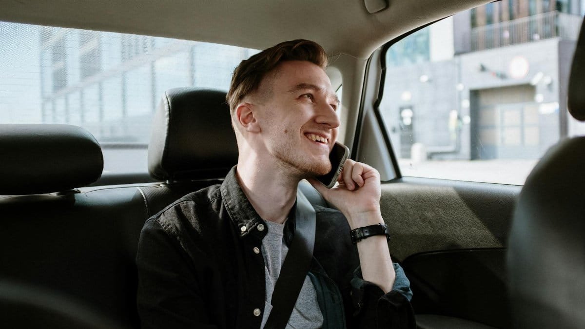 Smiling young man enjoying a phone call while seated in the back seat of a car during a daytime ride.