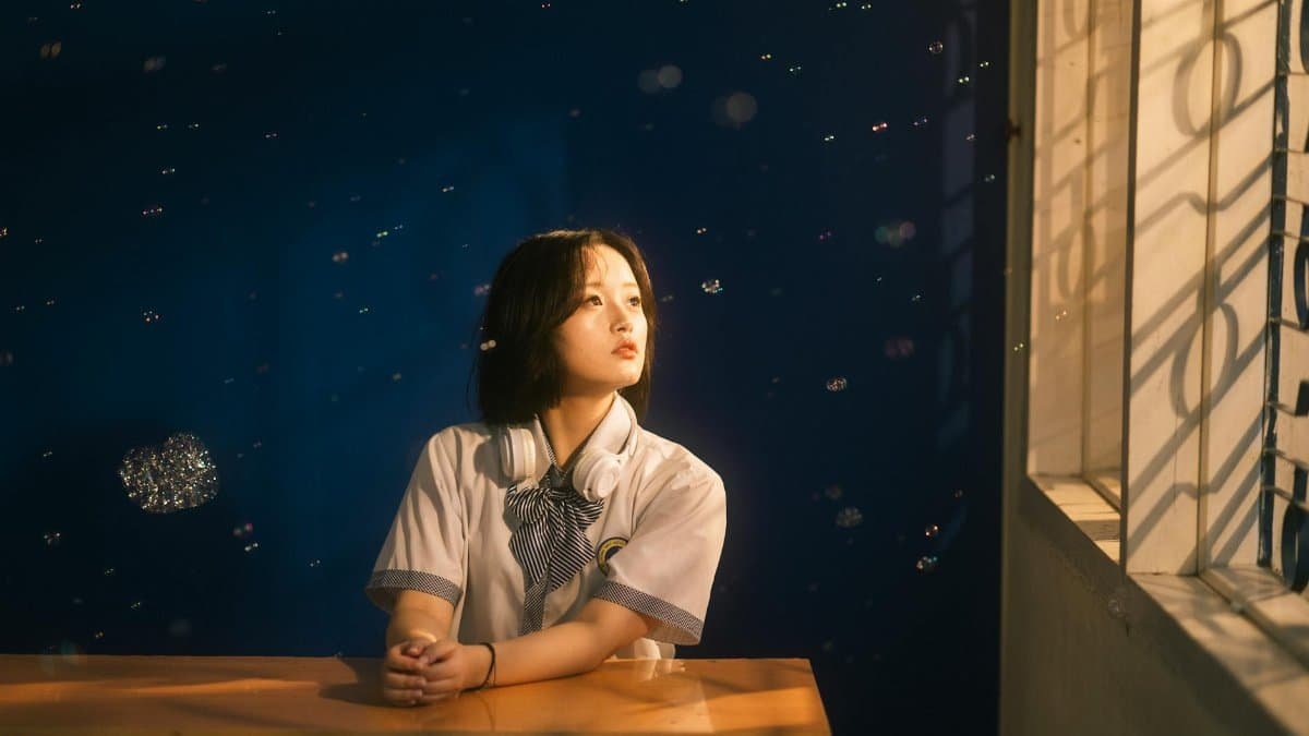 A student sits at a desk, gazing thoughtfully through a window, with gentle light and floating bubbles enhancing the serene atmosphere.