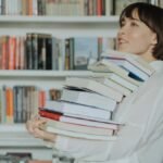 A woman joyfully holds a pile of books in a library, embodying the love of reading and learning.