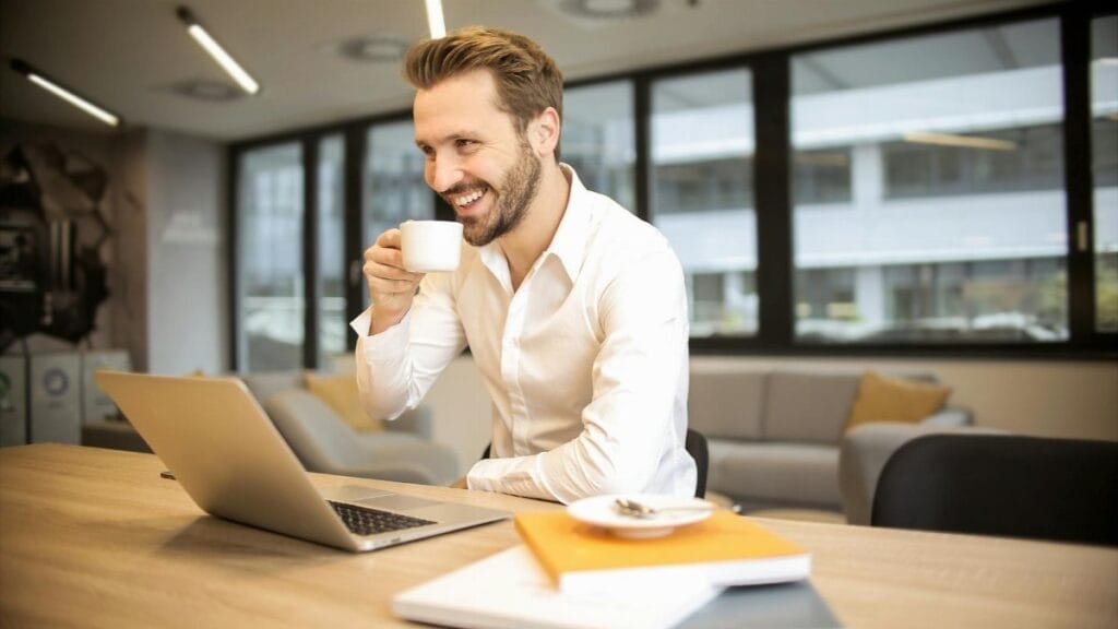 Man enjoying a coffee break while working in a modern office with laptop and books.