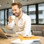 Man enjoying a coffee break while working in a modern office with laptop and books.