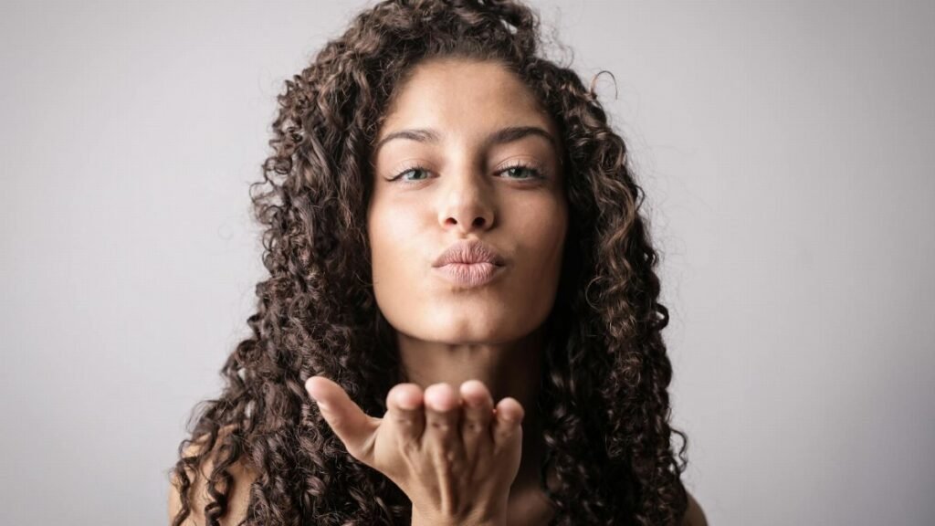 Elegant young woman with curly hair sending a playful kiss in studio. Perfect for beauty and lifestyle themes.