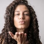 Elegant young woman with curly hair sending a playful kiss in studio. Perfect for beauty and lifestyle themes.