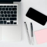 A top view of a modern workspace featuring a laptop, smartphone, pens, and a pink sticky note on a white desk.