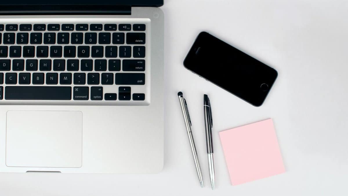 A top view of a modern workspace featuring a laptop, smartphone, pens, and a pink sticky note on a white desk.