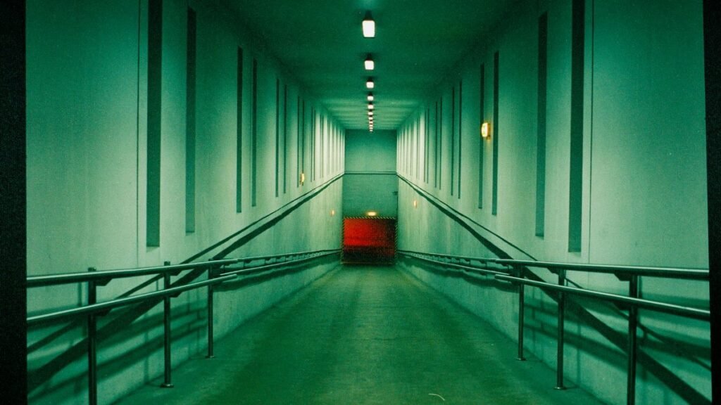 A long, empty corridor with dim lighting and an accessibility ramp leading to a red door.