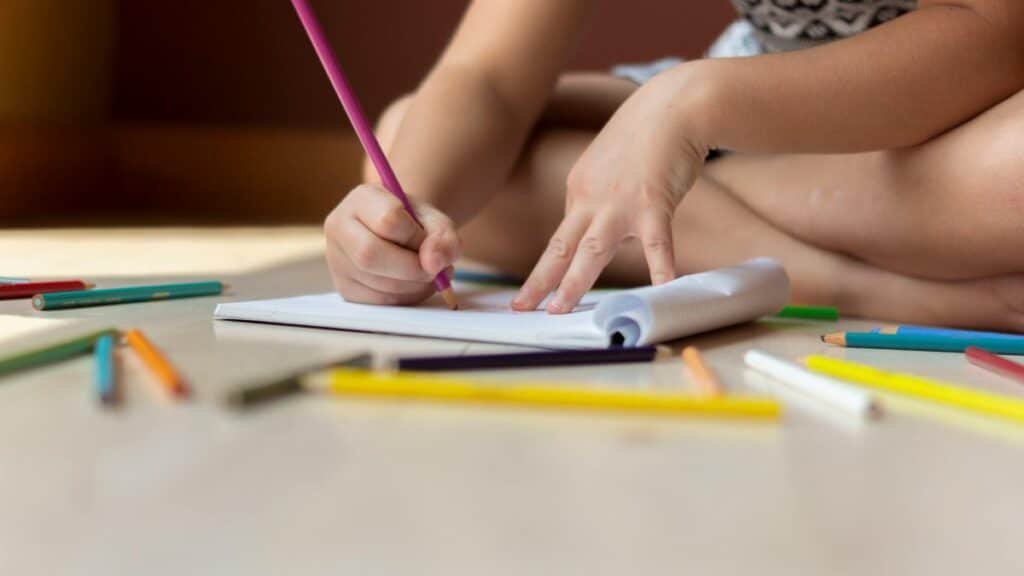 A child seated on the floor using colored pencils to draw on paper, showcasing creativity.