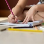 A child seated on the floor using colored pencils to draw on paper, showcasing creativity.