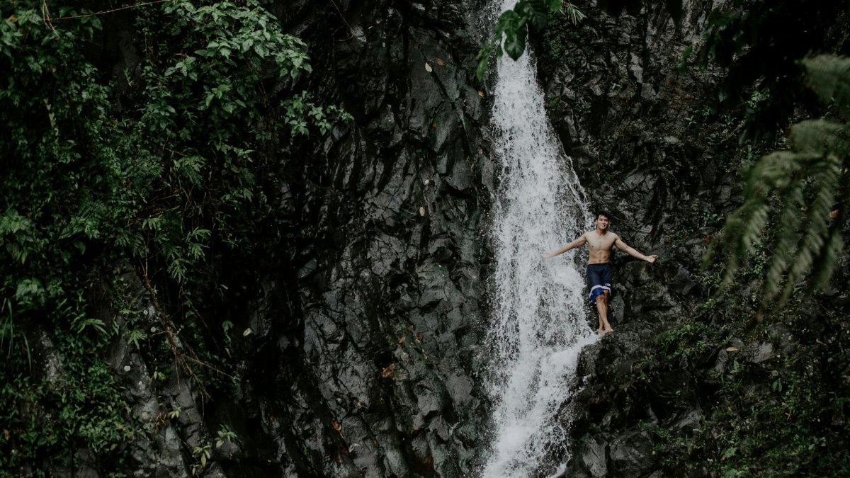 Unrecognizable male traveler in shorts standing on rough mountain near fast cascade in summer