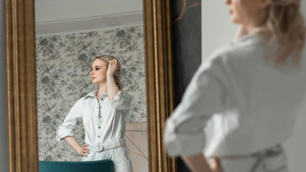 A fashionable woman poses confidently in a mirror reflection, exuding elegance indoors.