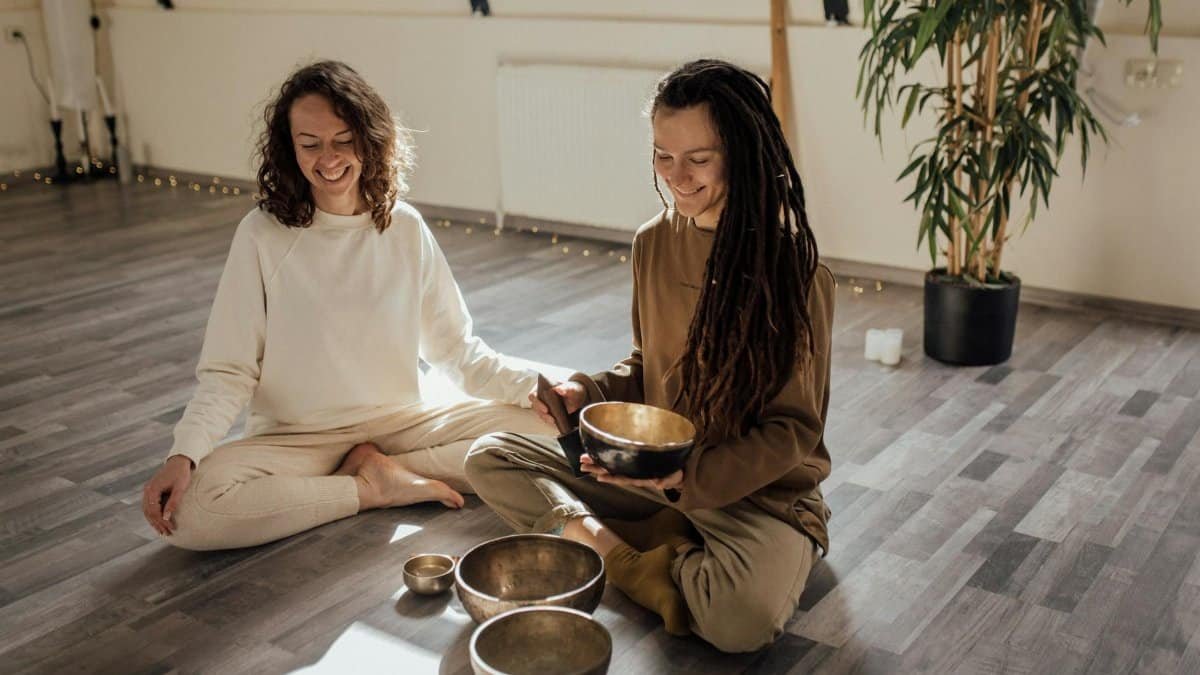 Two women sitting indoors with Tibetan singing bowls, engaging in a meditation session. Peaceful atmosphere.