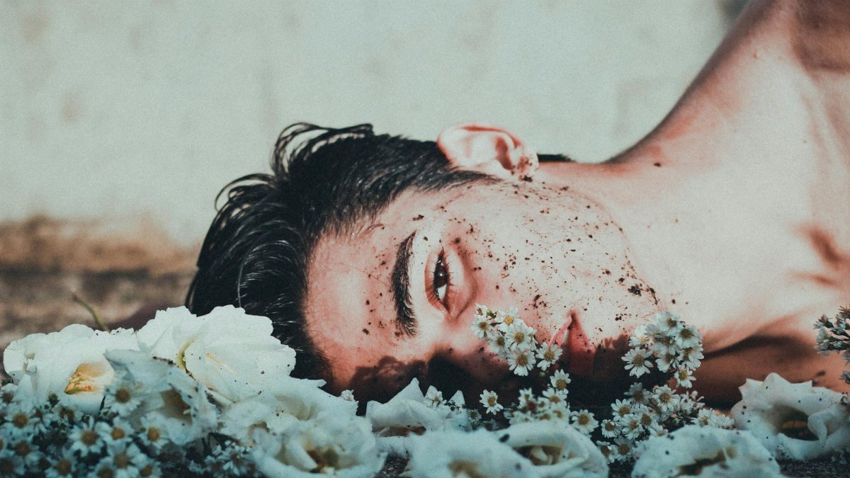 An ethereal portrait of a young adult male lying among flowers, conveying themes of vulnerability and mental health.