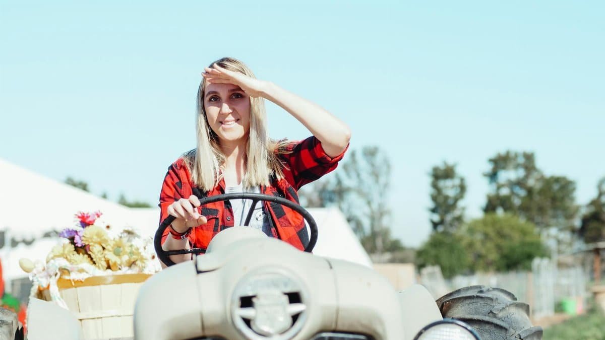 Cheerful woman driving a tractor outdoors, shielding eyes from the sun.
