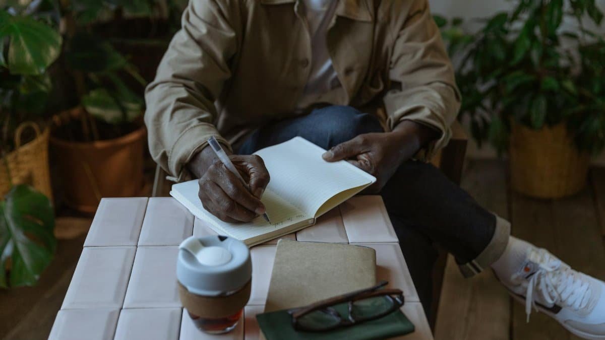 A man takes notes in a notebook while sitting indoors surrounded by plants, creating a cozy atmosphere.