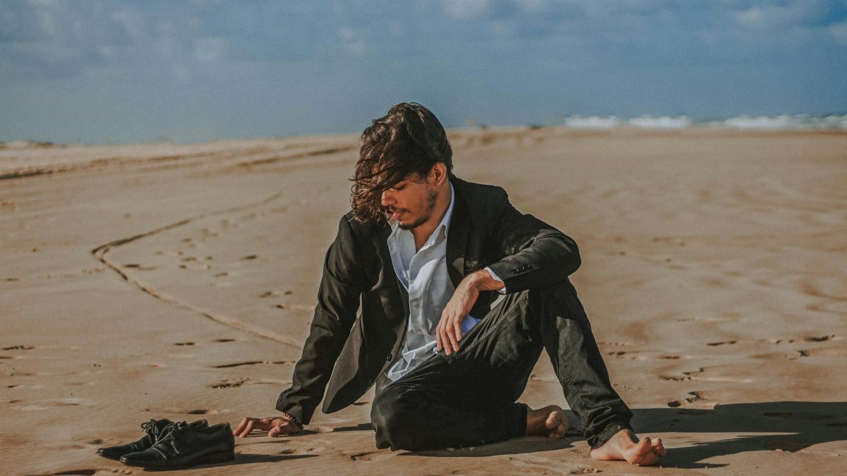 Man in suit sitting barefoot on a sandy beach, contemplating surroundings.