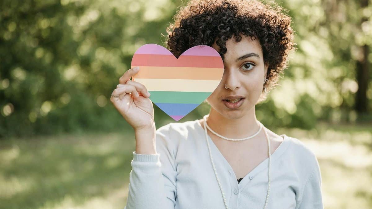 A woman stands outdoors holding a colorful rainbow heart, symbolizing pride and diversity.