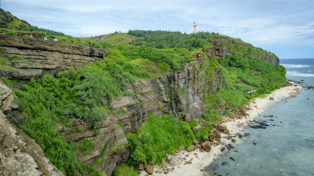 Coastal cliff with lush greenery and a sandy beach. Lighthouse visible on the hilltop.