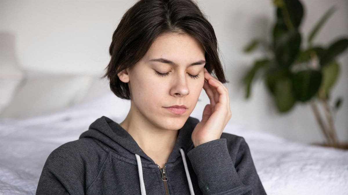 A young woman with closed eyes sits thoughtfully indoors, wearing a hoodie, suggesting deep reflection or meditation.