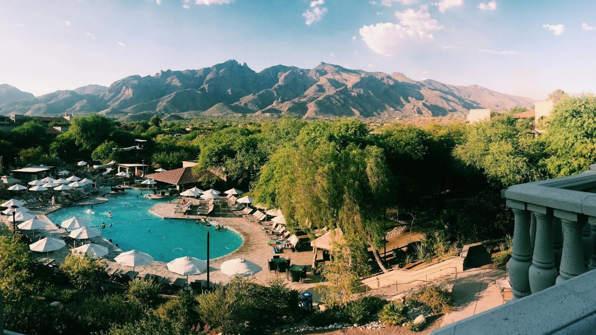 Aerial view of a resort pool surrounded by trees, with mountains in the background under a sunny sky.