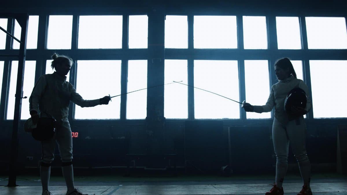 Two female fencers in protective gear face off in an indoor training setting.