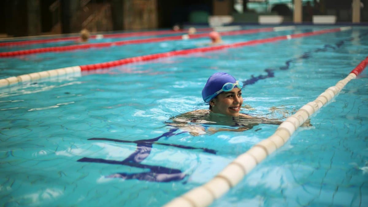 Woman enjoying a swim in an indoor pool with a blue cap and goggles, exuding joy and relaxation.