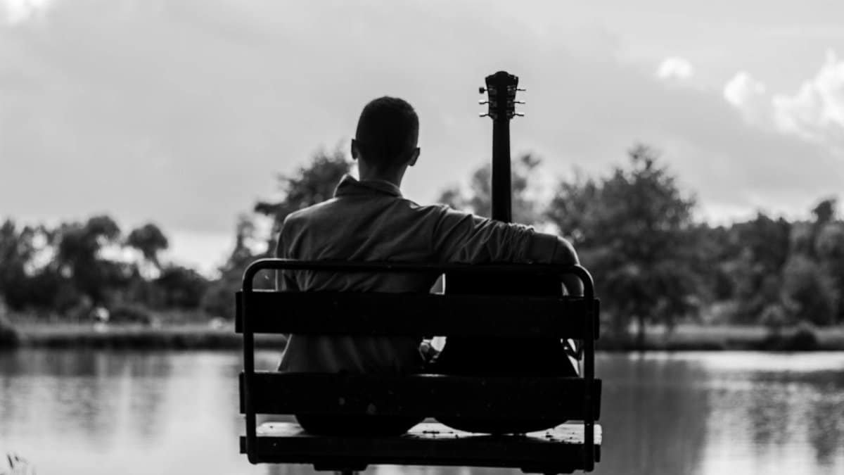 A serene silhouette of a guitarist sitting on a bench by a lake at dusk, in monochrome.