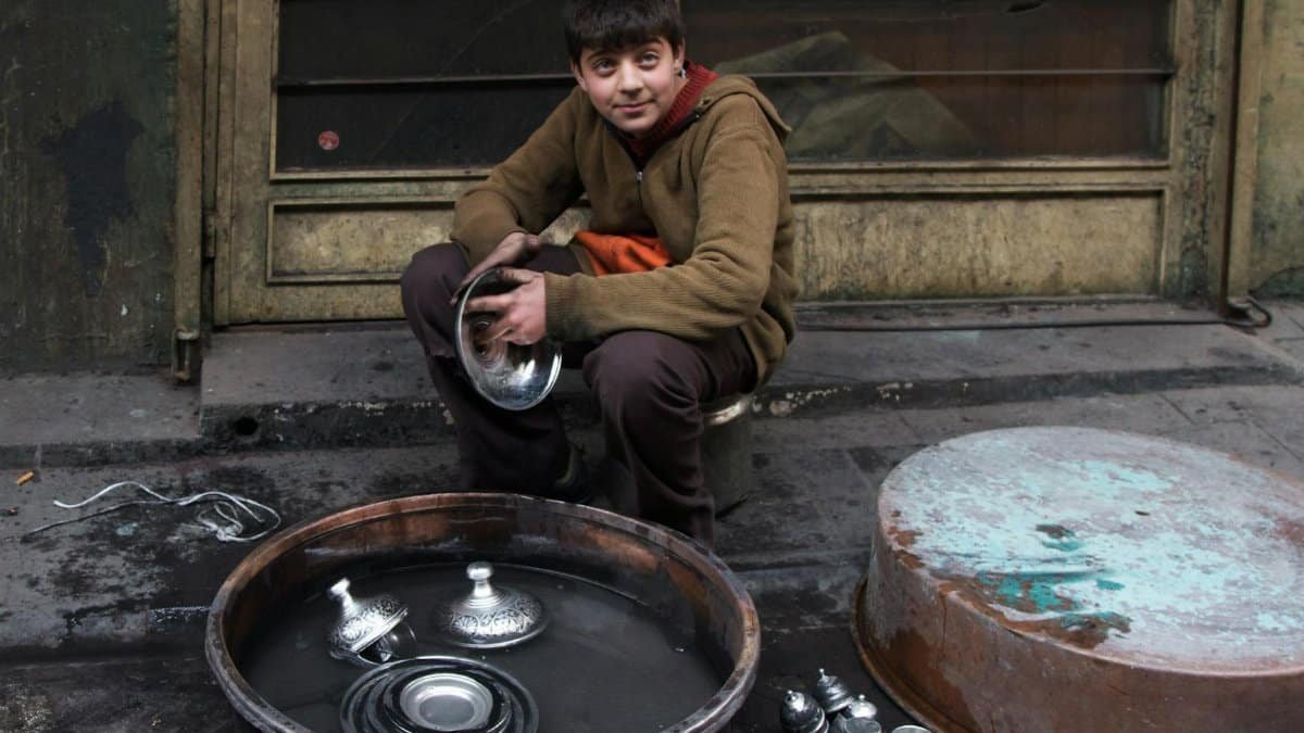 A teenager sits on a stool washing metal dishes on an urban street, surrounded by various utensils and a large basin.