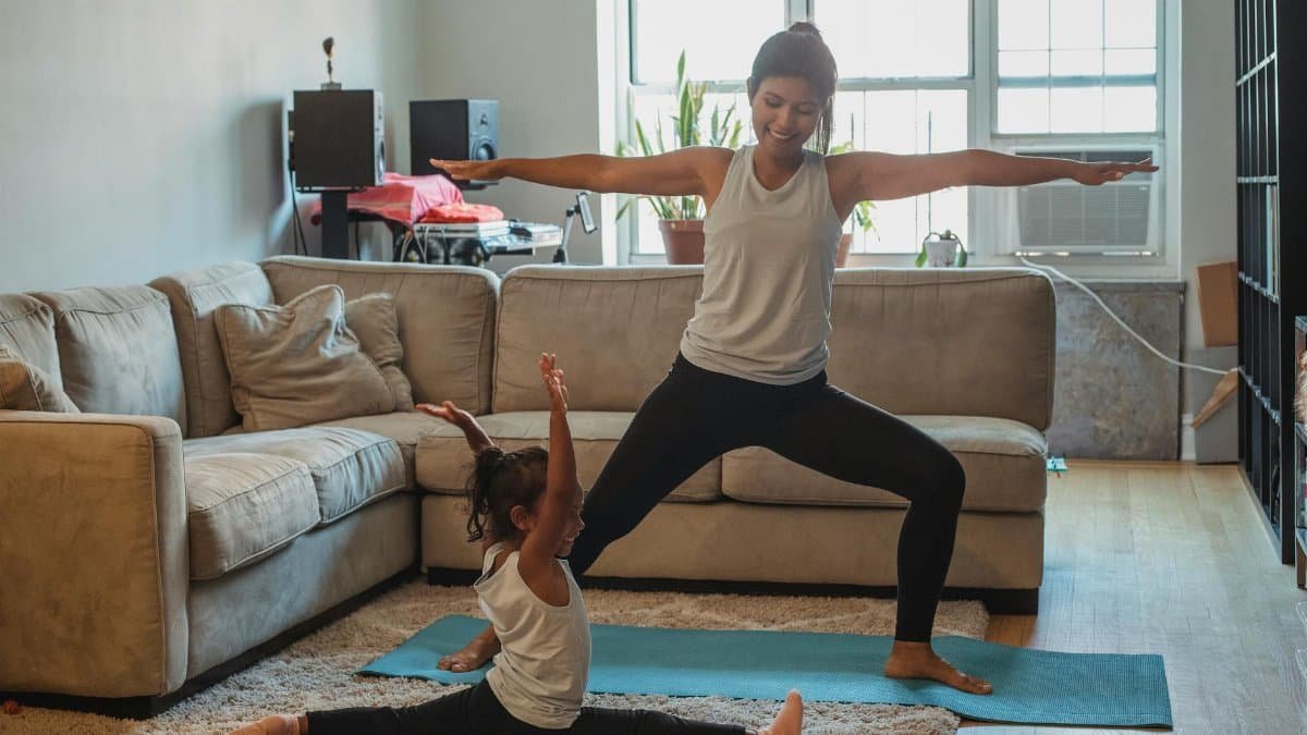 Full body cheerful ethnic mother in sportswear practicing yoga and performing Warrior Pose on mat near cute daughter doing splits in light living room