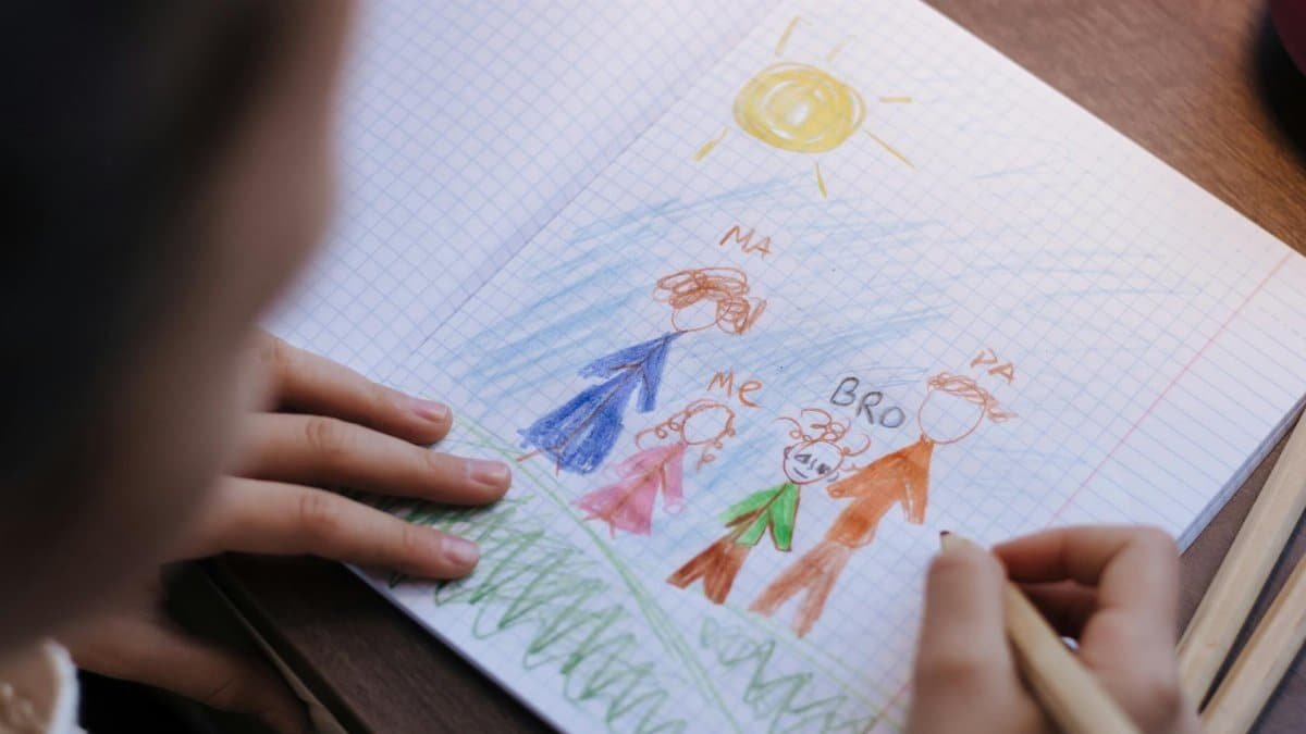 A child drawing a family portrait with crayons in a notebook on a wooden table.