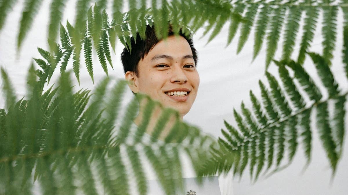 A smiling man photographed through lush green ferns, creating a vibrant, happy portrait in nature.