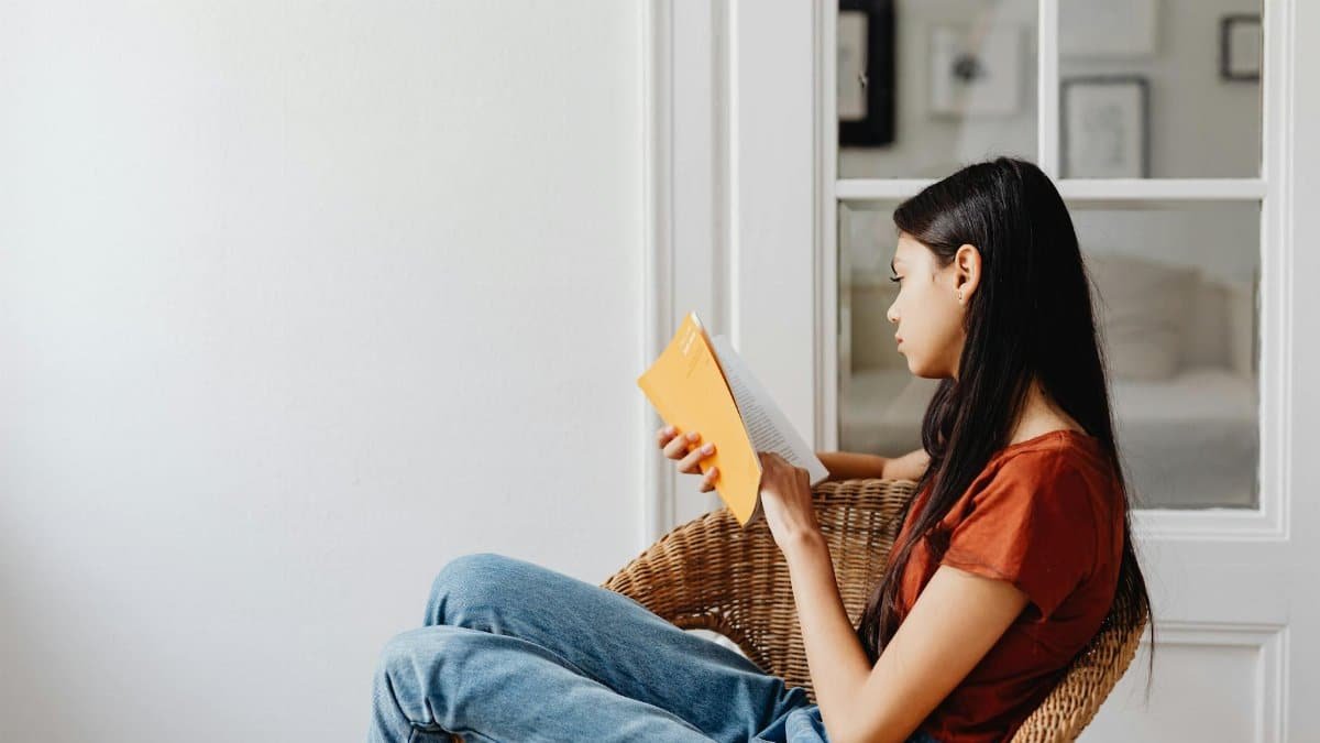 A young woman sits in a woven chair reading a book in a bright room.