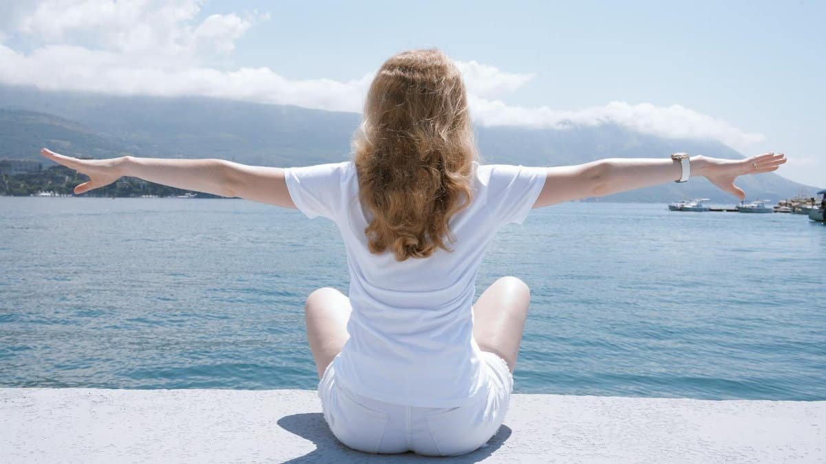 Woman sitting with open arms by the water, embracing nature and serenity.